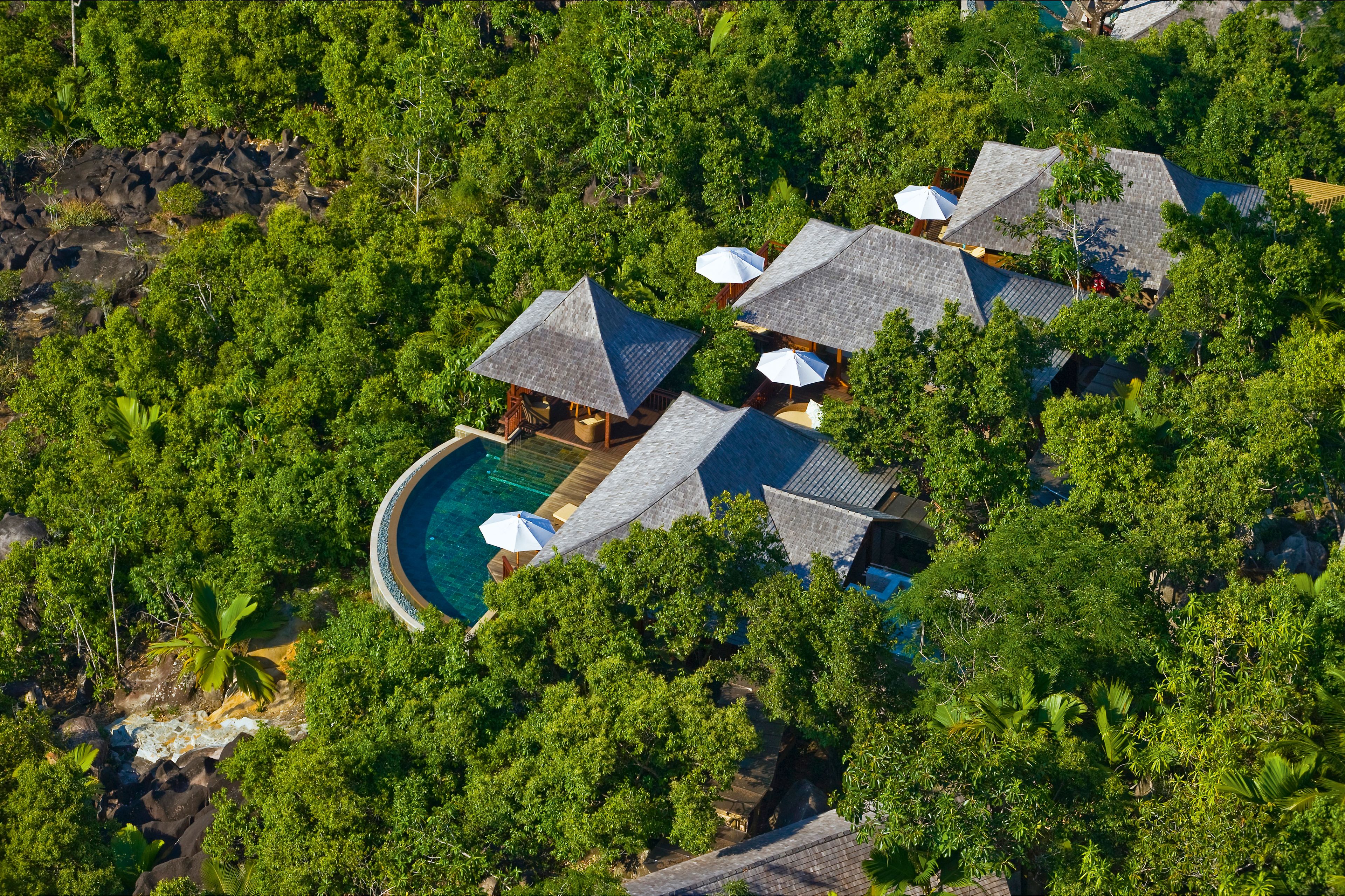 An aerial view of a resort with multiple thatched-roof cottages, a central swimming pool, and lush surrounding vegetation.