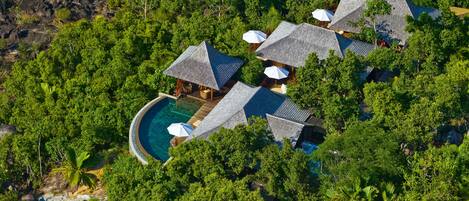 An aerial view of a resort with multiple thatched-roof cottages, a central swimming pool, and lush surrounding vegetation.