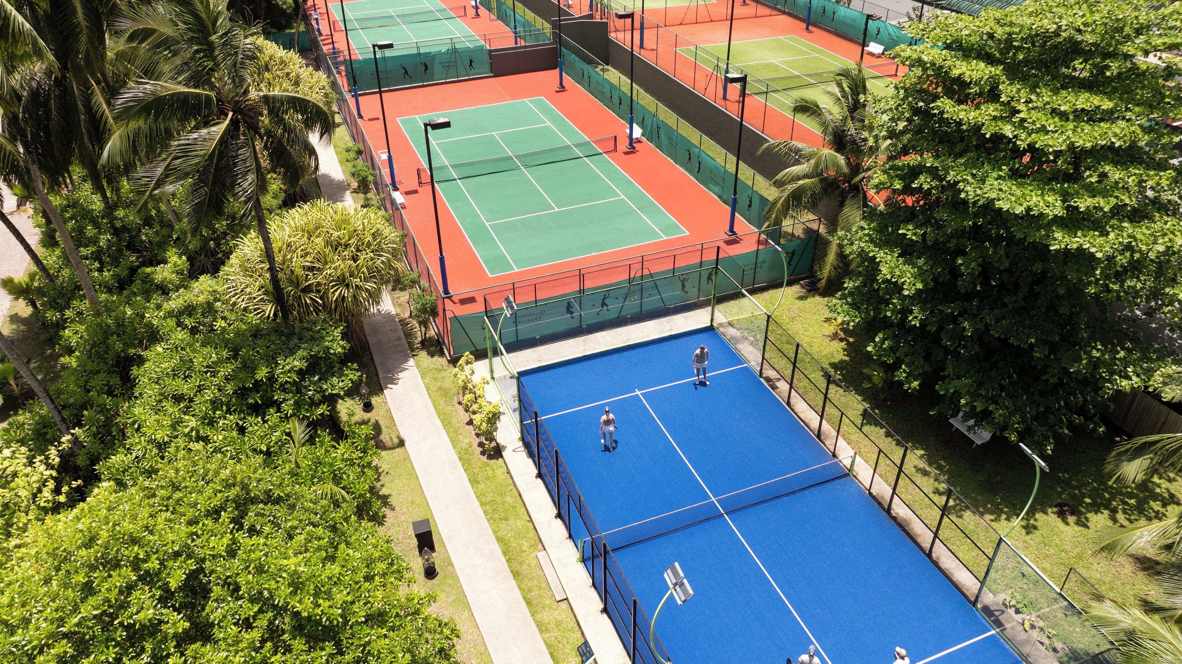 Ein Luftbild eines Tennisplatzes mit mehreren Spielfeldern, umgeben von üppiger Vegetation und Palmen.