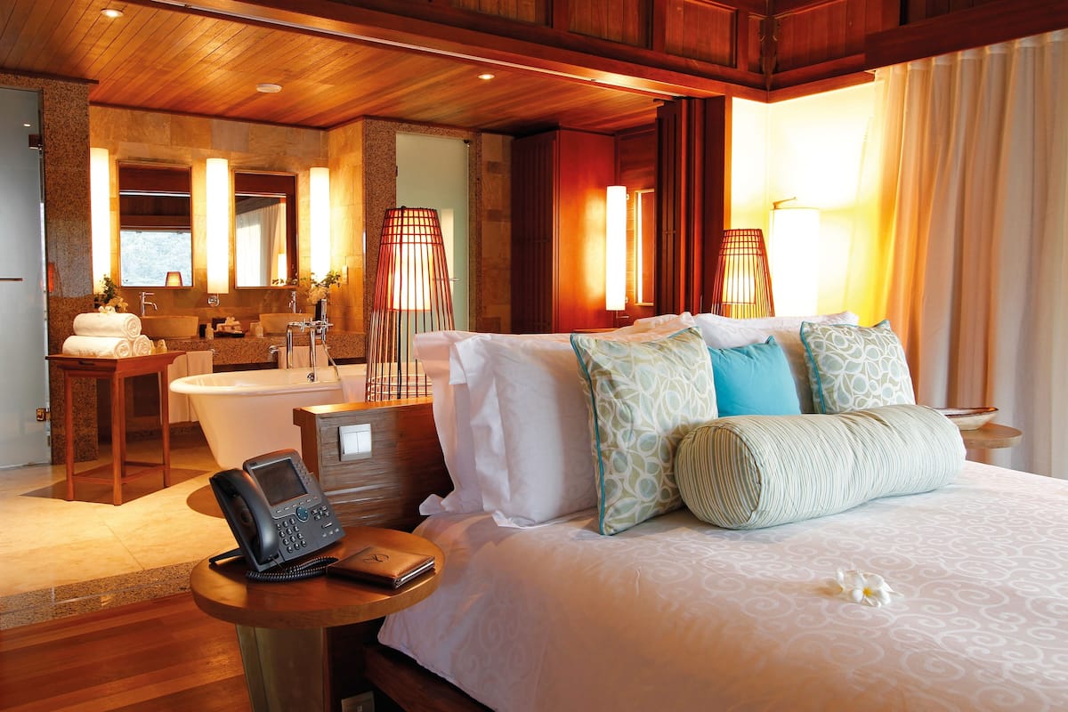 Habitación de hotel con techo de madera, una cama grande con varias almohadas, una mesita de noche con un teléfono y una billetera, un baño con bañera y lavamanos, y un espejo.