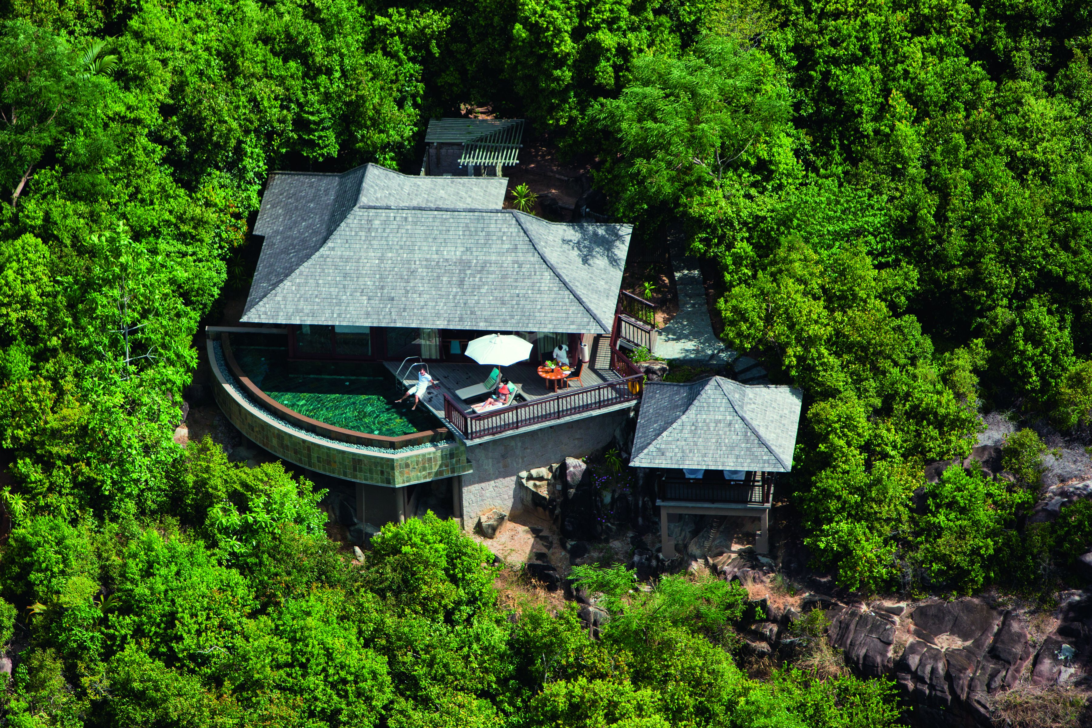 A large, multi-story building surrounded by dense forest, featuring a rooftop terrace with outdoor seating and a small adjacent structure.