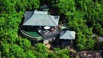A large, multi-story building surrounded by dense forest, featuring a rooftop terrace with outdoor seating and a small adjacent structure.
