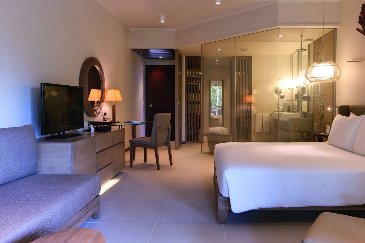 Habitación de hotel moderna con cama, sofá, televisor, escritorio y baño visible a través de una puerta de cristal.