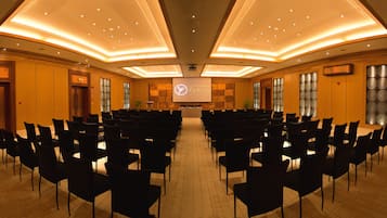 Une salle de conférence avec des rangées de chaises disposées face à un écran de présentation, des panneaux de bois et un sol carrelé.