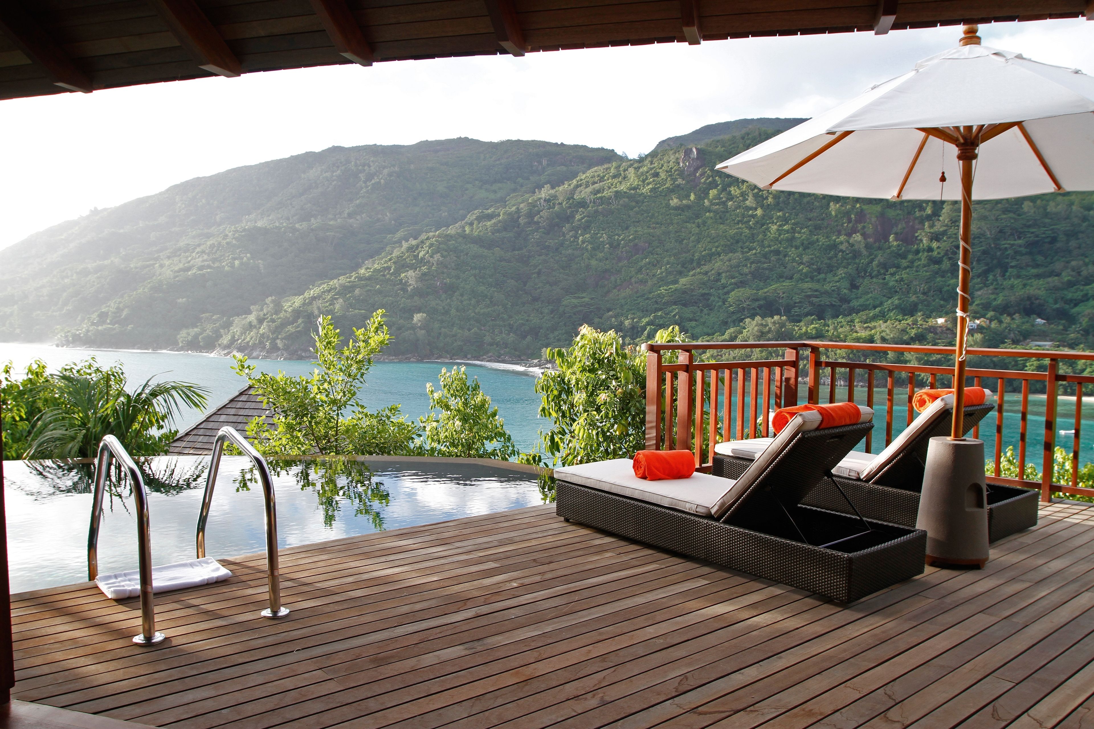 Um deck de madeira com piscina, espreguiçadeiras e um guarda-sol, com vista para uma paisagem montanhosa e um corpo de água.