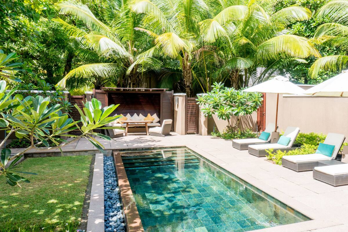 Una piscina con sillas de descanso y sombrillas, rodeada de abundante vegetación y palmeras.