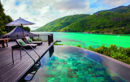 A wooden deck with lounge chairs and an umbrella overlooking a turquoise pool with a clear view of a mountain and a body of water.