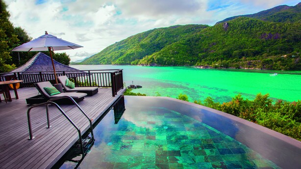 A wooden deck with lounge chairs and an umbrella overlooking a turquoise pool with a clear view of a mountain and a body of water. - Constance Ephélia (Mahé Island)