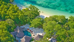 An aerial view of a tropical resort with a clear turquoise sea, lush greenery, and a swimming pool area with thatched-roof huts.