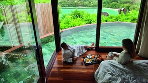 A bedroom with a large glass window overlooking a lush garden and a pool area. - Constance Ephélia (Mahé Island)