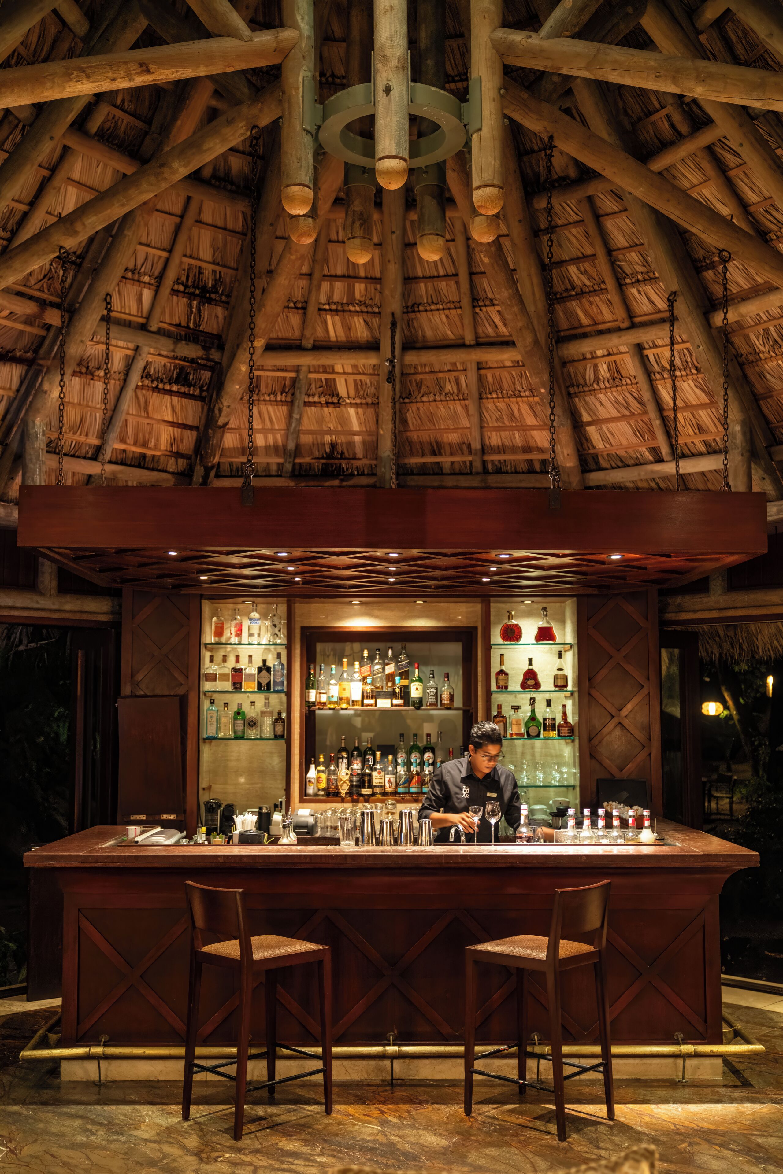 Un bar avec un toit de chaume, des poutres en bois et une variété de bouteilles et de verres exposés sur des étagères.