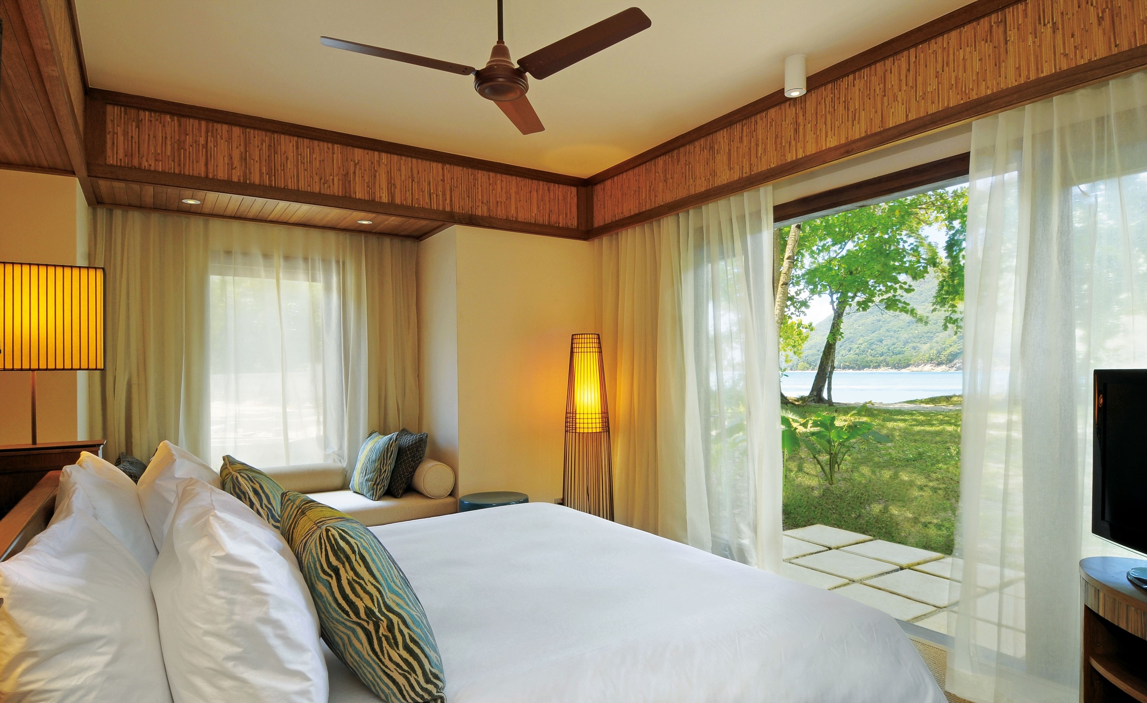 A hotel room with a bed, a ceiling fan, a lamp, and a view of a natural landscape through large windows.