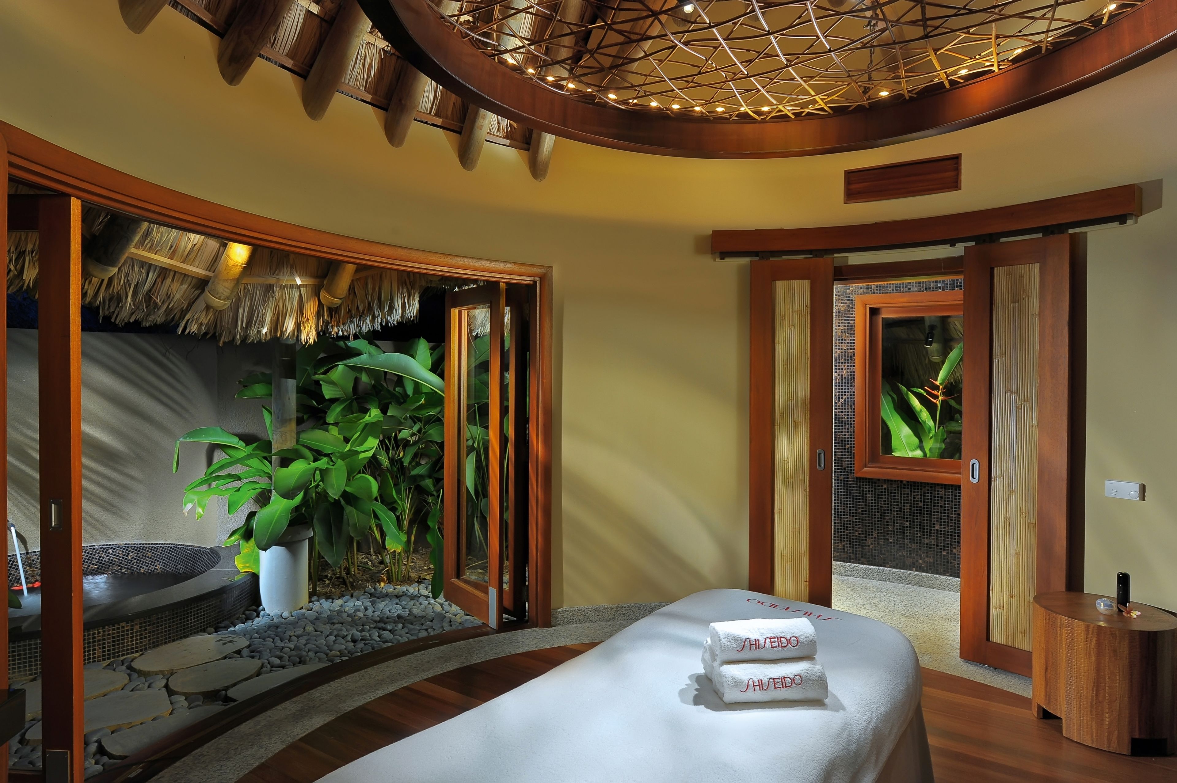 Ein Spa-Raum mit einem Bett, einer großen Kübelpflanze, einer kleinen Innenwassereinrichtung und Gartenblick durch eine Glastür.