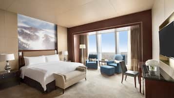 A hotel room with a large bed, a desk with a TV, a seating area with chairs, and a large window with a view.