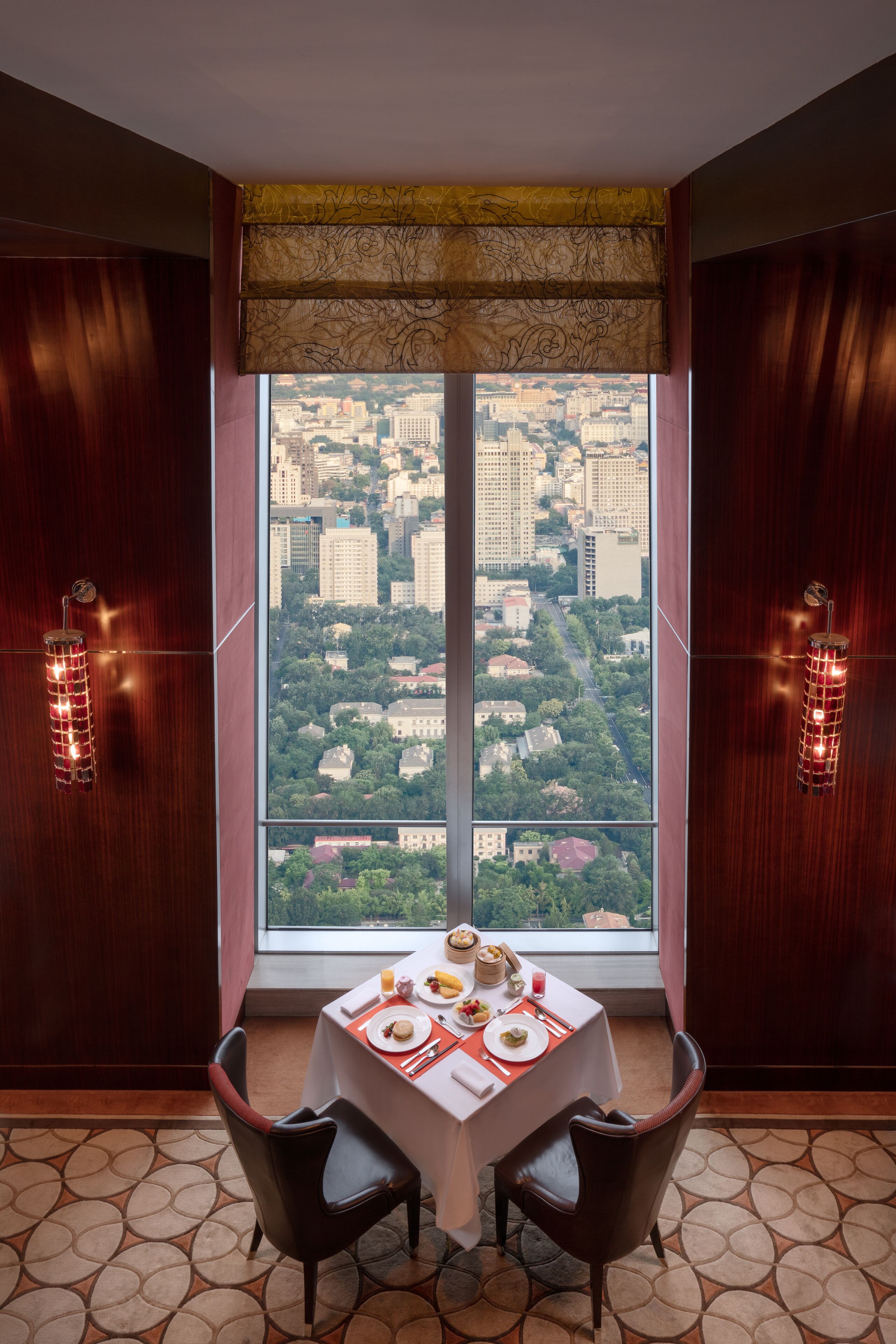 A dining area with a table set for two, a city view through large windows, and a patterned floor.