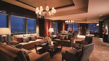 Un vaste salon comprenant un grand canapé, des fauteuils et une table basse. Un lustre est suspendu au plafond et une grande fenêtre offre une vue sur la ville.