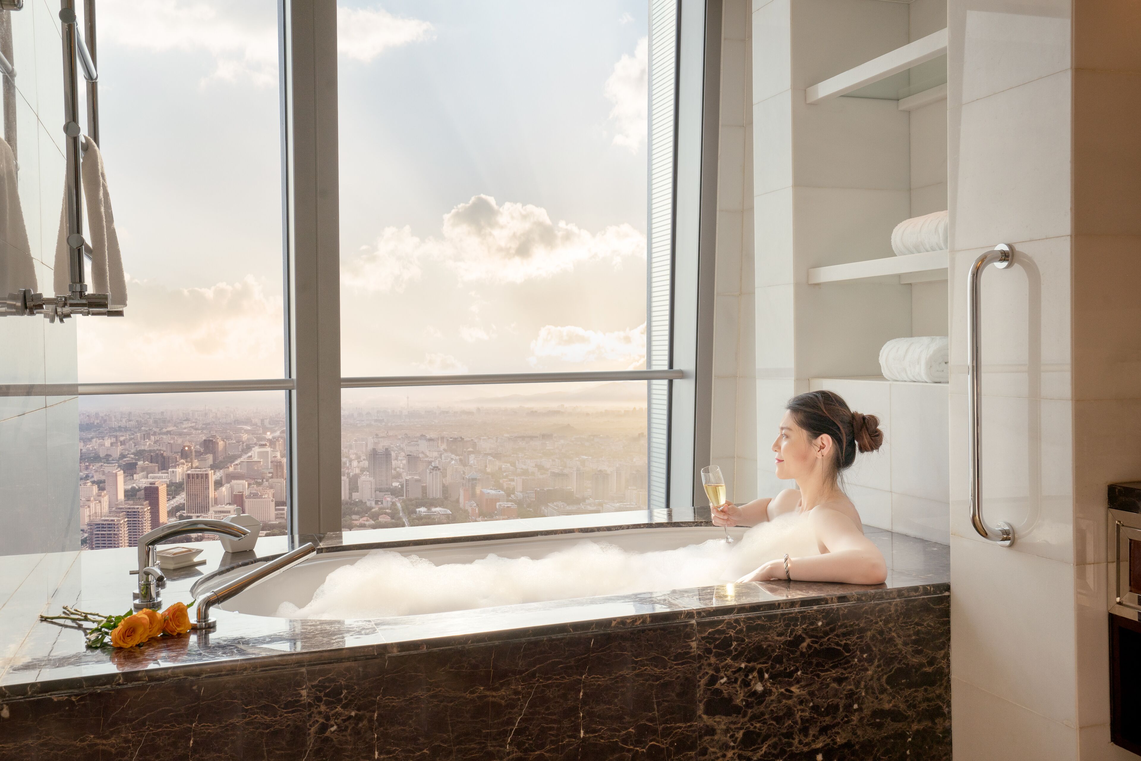 Une femme se détend dans une baignoire, avec vue sur la ville, un verre de champagne à la main et une rose posée sur le bord de la baignoire.