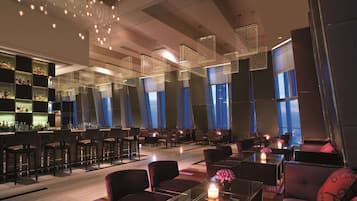 A modern bar area with high tables, bar stools, and a variety of bottles. There are multiple seating areas with tables and chairs, and large windows providing views.