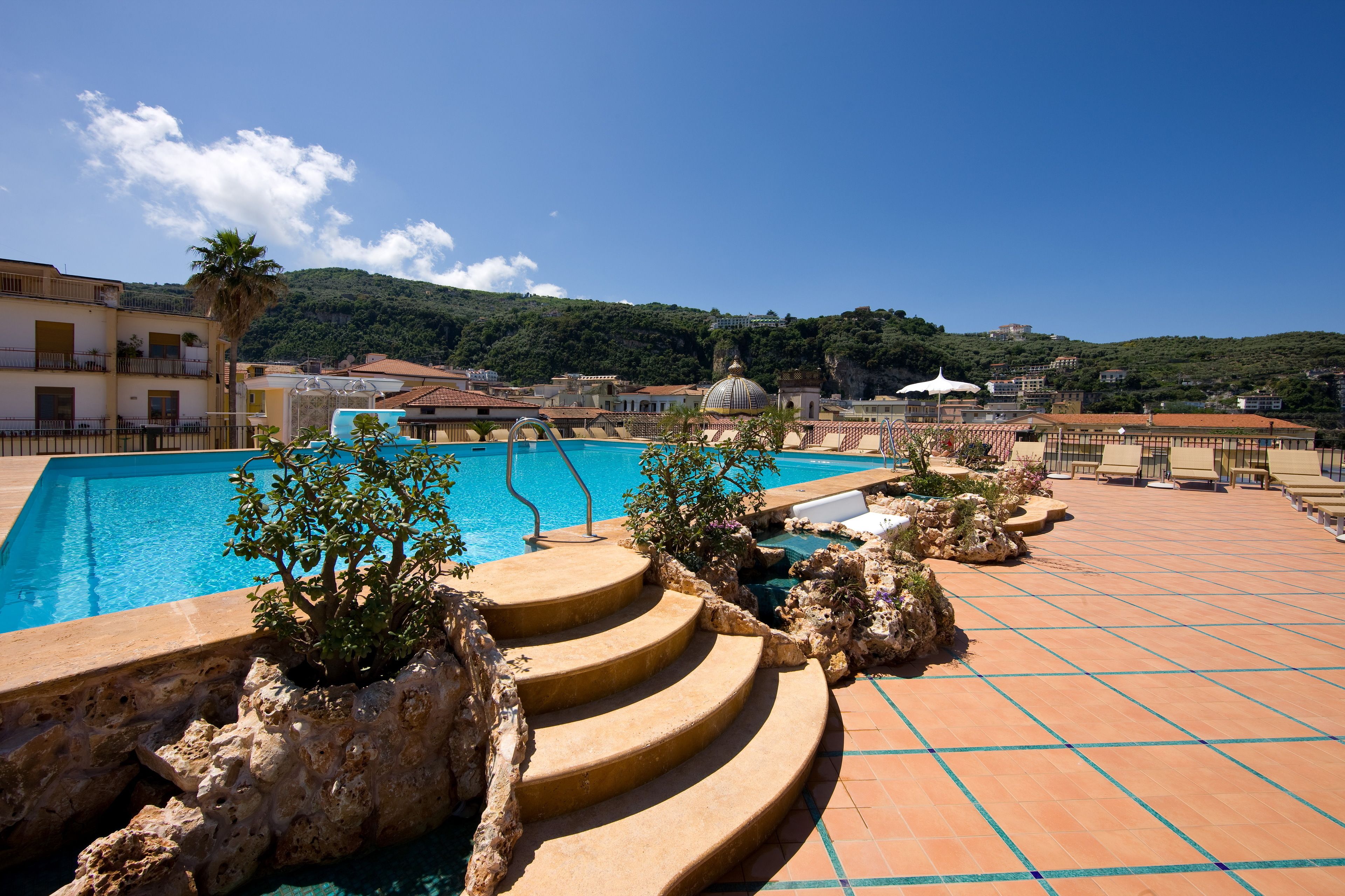 Una piscina amb escales d'accés, envoltada per un solàrium amb rajoles i algunes plantes.