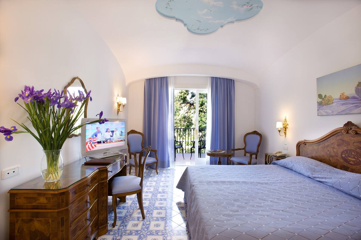A hotel room with a bed, a desk with a computer, a chair, a vase with flowers, and a view of the outdoors.