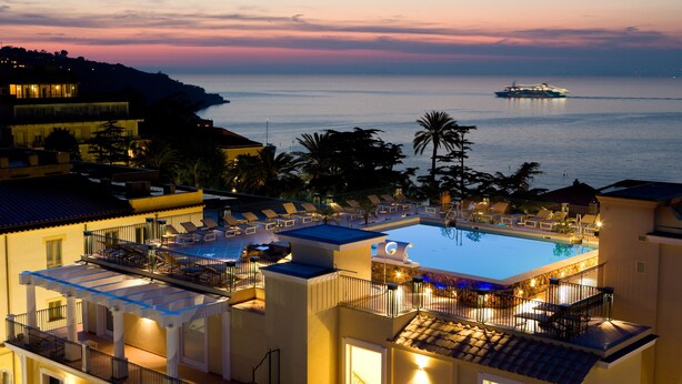 Un complex turístic amb piscina, gandules i vistes al mar al capvespre.