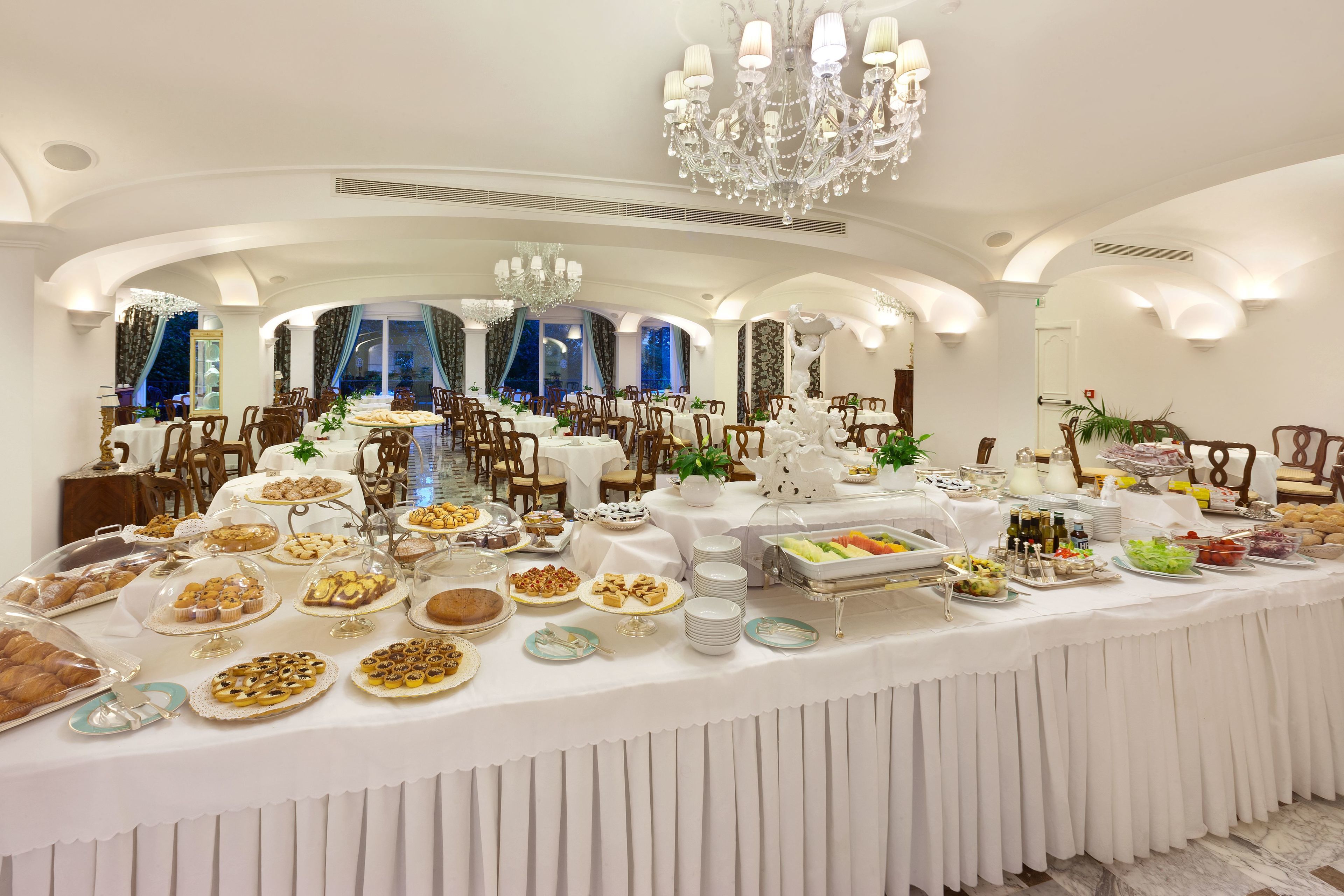 A buffet setup in a well-lit dining area with multiple tables and chairs, elegant chandeliers, and large windows.