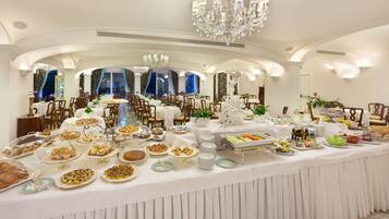A buffet setup in a well-lit dining area with multiple tables and chairs, elegant chandeliers, and large windows.