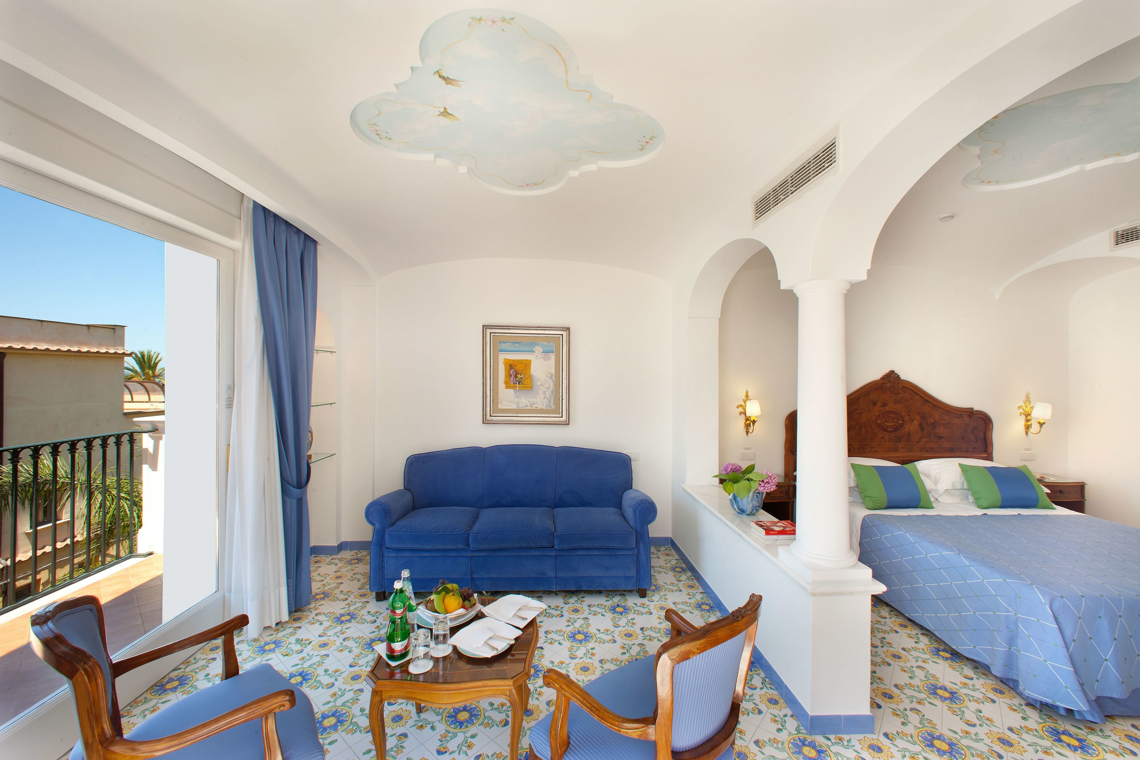 Quarto de hotel com varanda, um sofá azul, uma mesa de madeira com uma garrafa e copos, uma cama com roupa de cama azul e um quadro na parede.