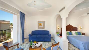 Quarto de hotel com varanda, um sofá azul, uma mesa de madeira com uma garrafa e copos, uma cama com roupa de cama azul e um quadro na parede.