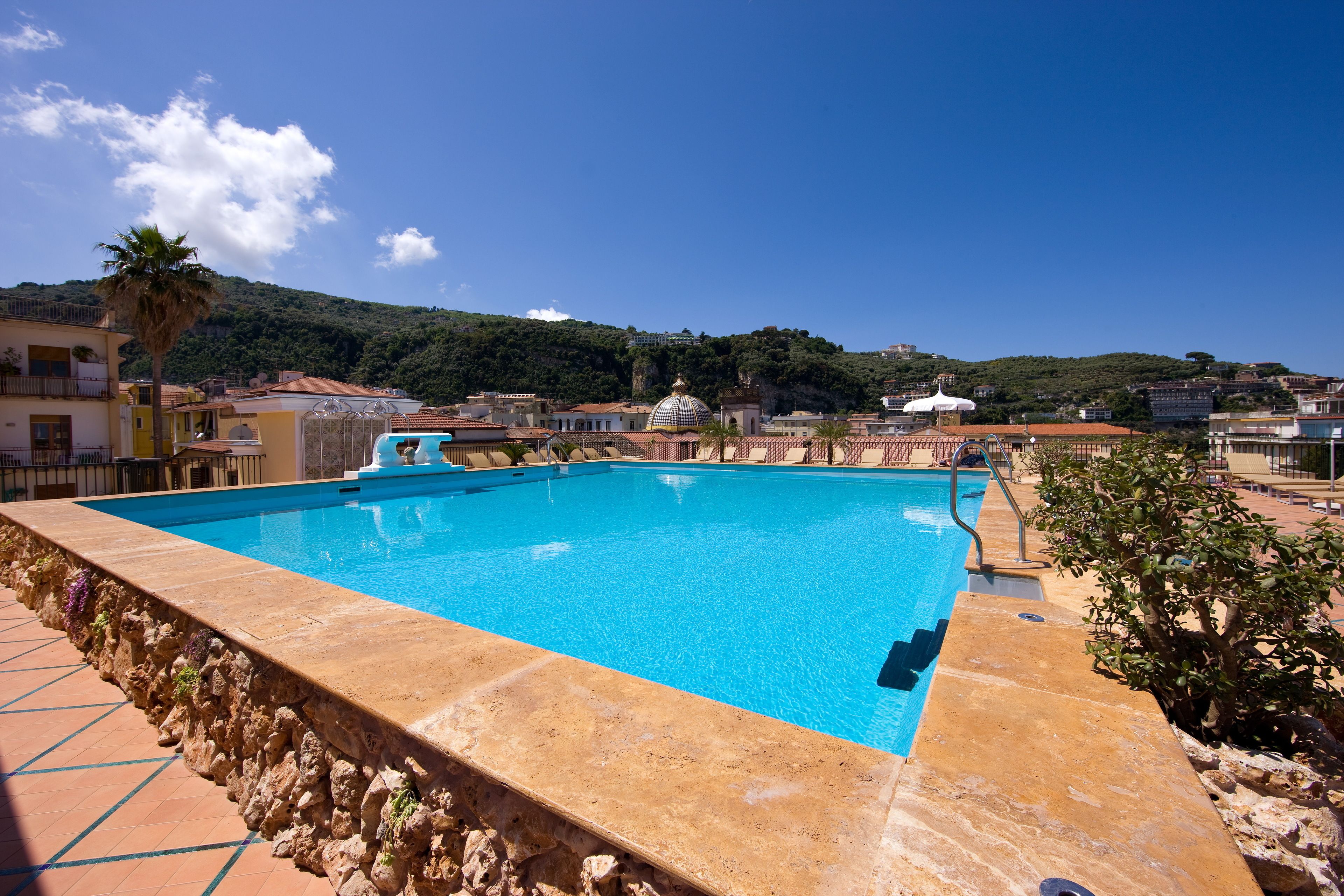 Una piscina con bordo in pietra, circondata da edifici e una collina sullo sfondo.