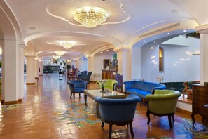 A spacious lobby with a high ceiling, ornate chandeliers, and a tiled floor with a floral pattern. There are several seating areas with upholstered chairs and a sofa.