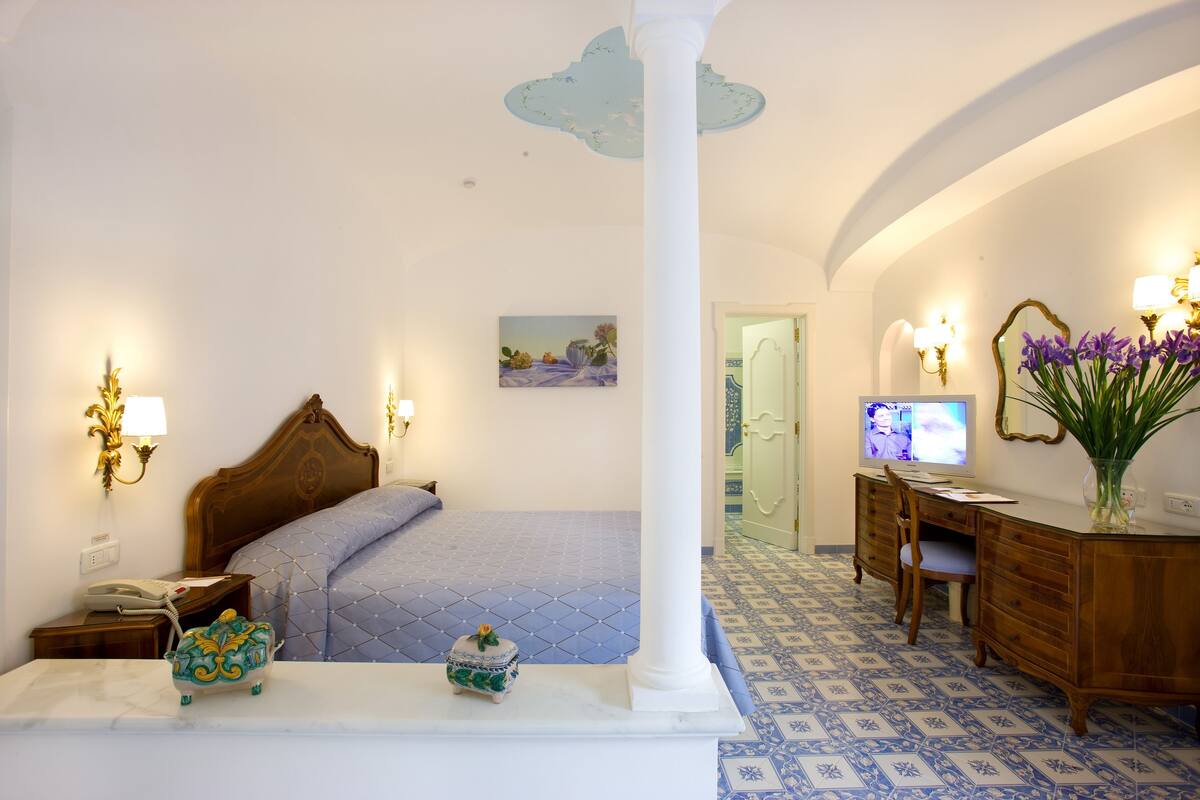 A hotel room with a bed, a desk, a chair, a television, and a vase with flowers.