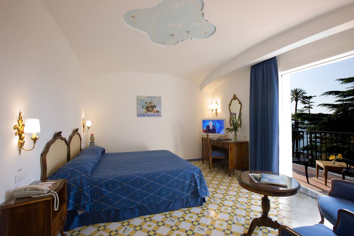 A hotel room with a bed, a desk, a chair, a small table, and a balcony with a view of palm trees.