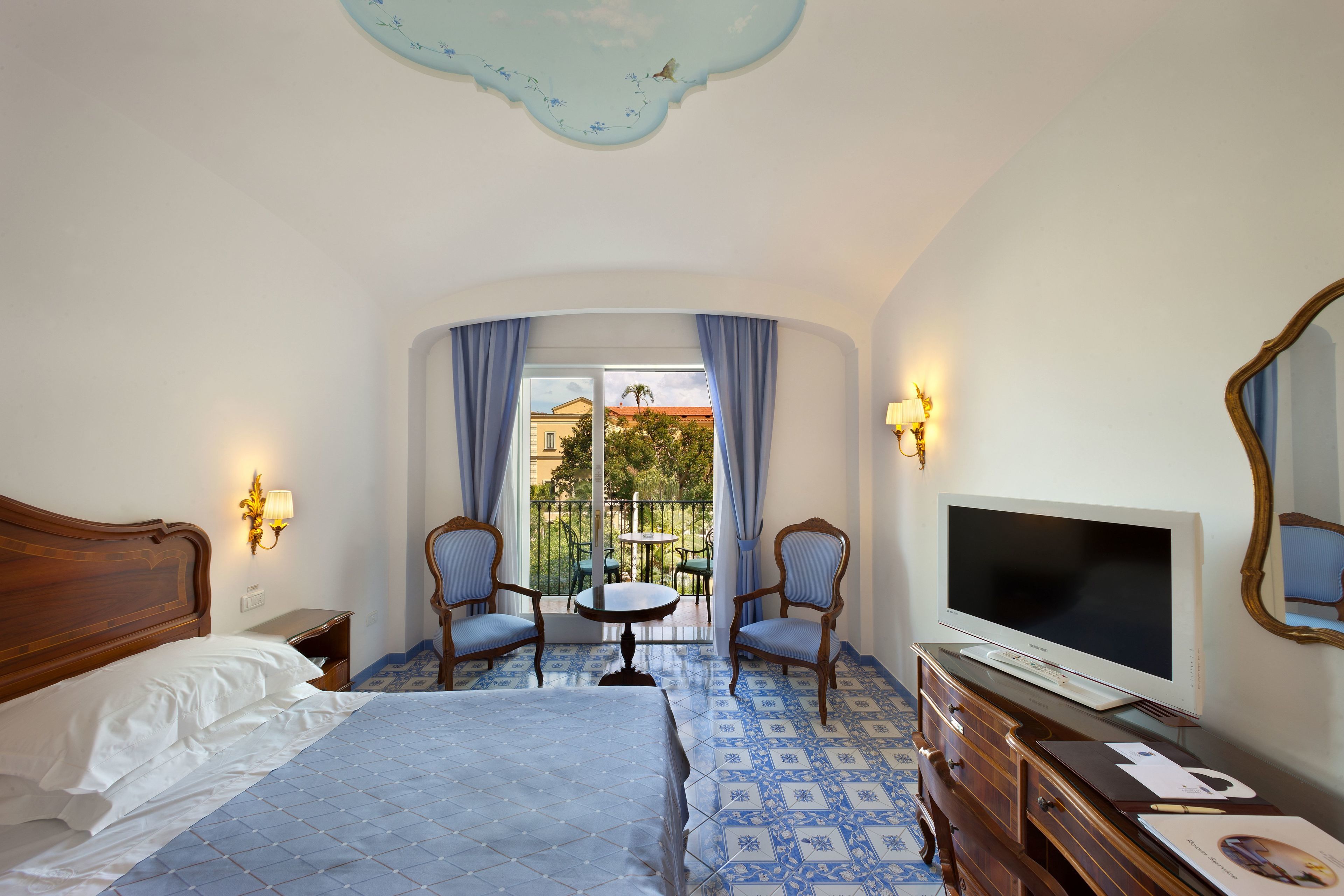 A hotel room with a bed, a TV, a dresser, and a balcony with a table and chairs.
