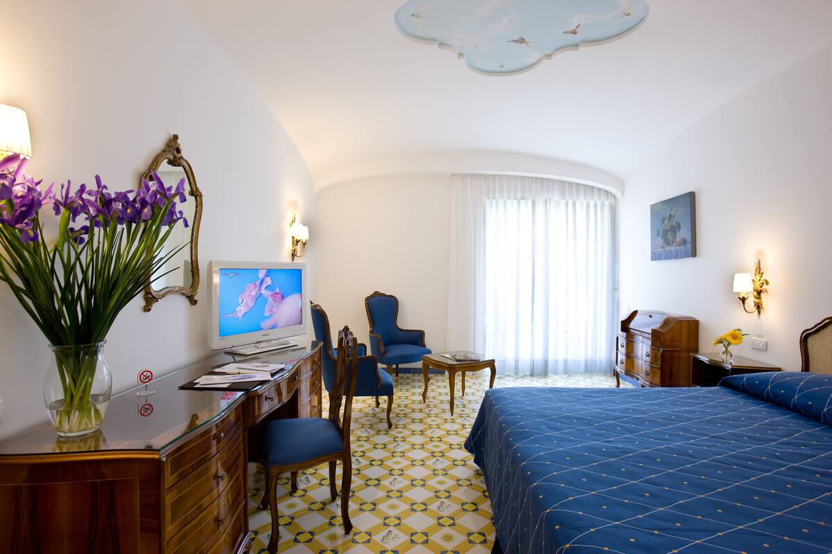 A hotel room with a bed, a desk with a computer, a chair, a small table, and a vase with flowers.