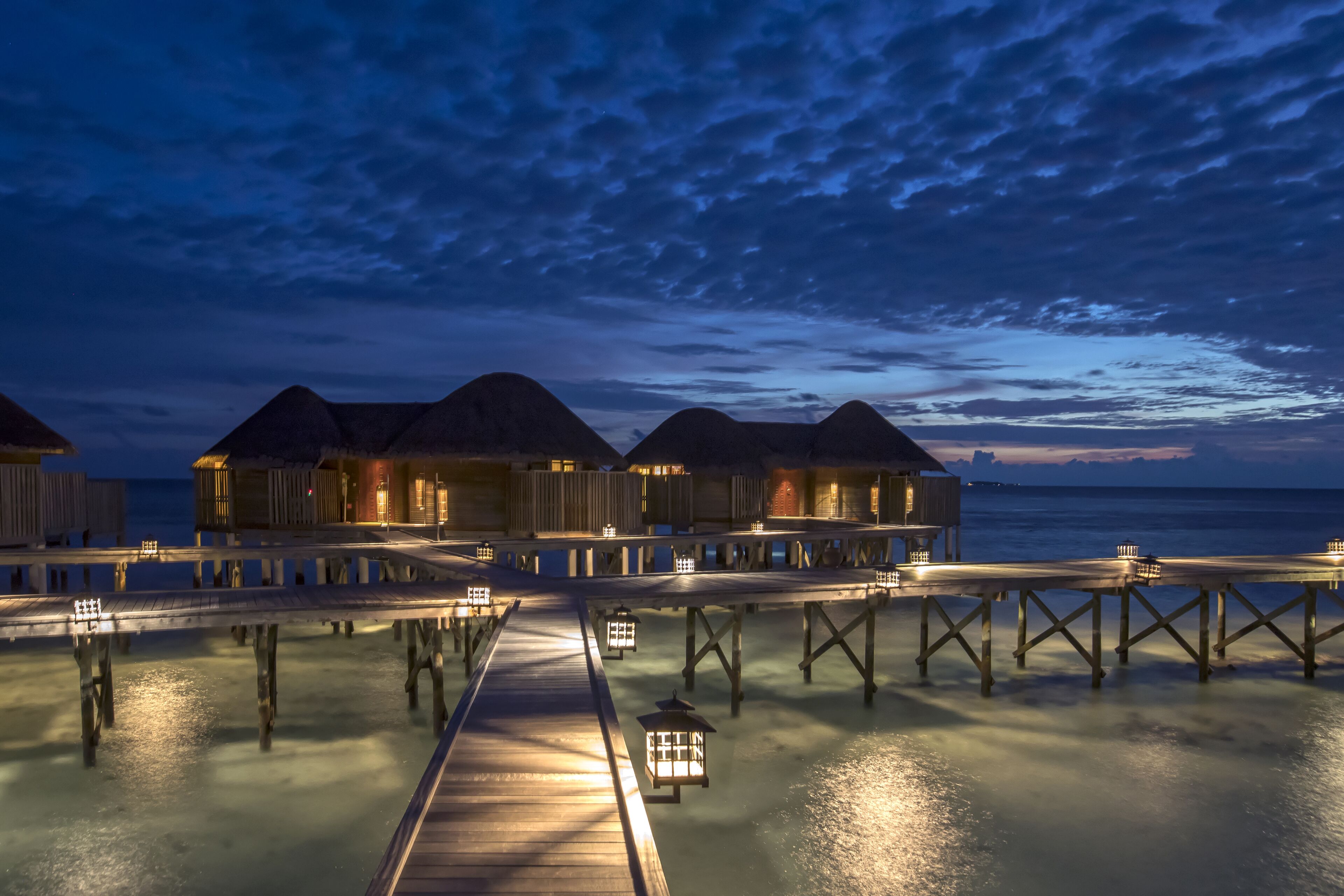 Ein Holz-Overwater-Resort mit strohgedeckten Bungalows, die durch einen Holzsteg über ruhigem Wasser verbunden sind.