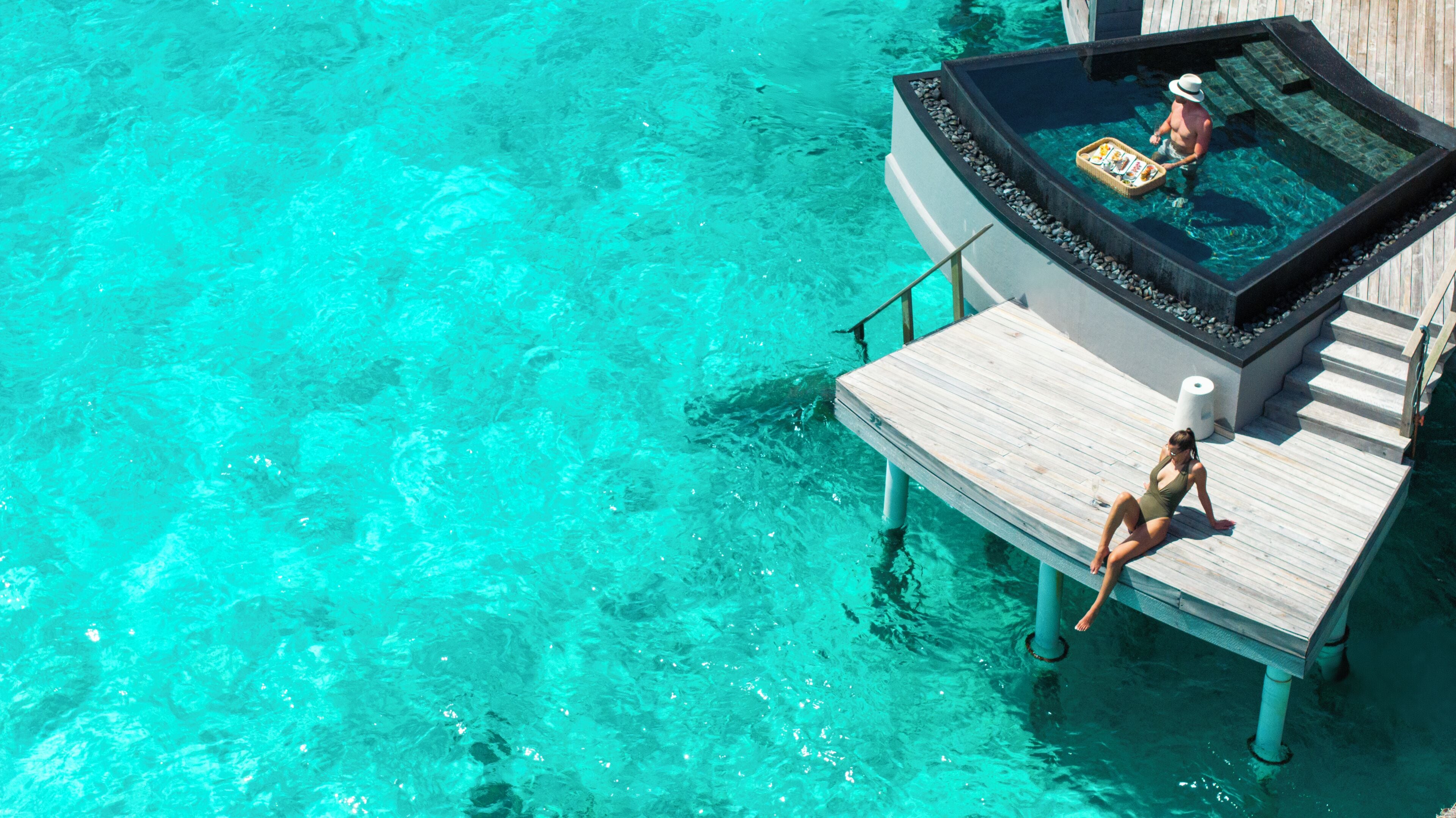 Une terrasse en bois s’étendant au-dessus d’une eau turquoise, un jacuzzi où se trouve une personne, et une femme assise sur la terrasse.
