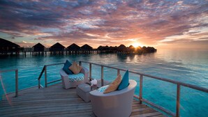 A wooden deck with two wicker lounge chairs, one with a blue cushion and the other with a beige cushion, overlooking a turquoise sea with overwater bungalows in the background. - Constance Halaveli (Halaveli)