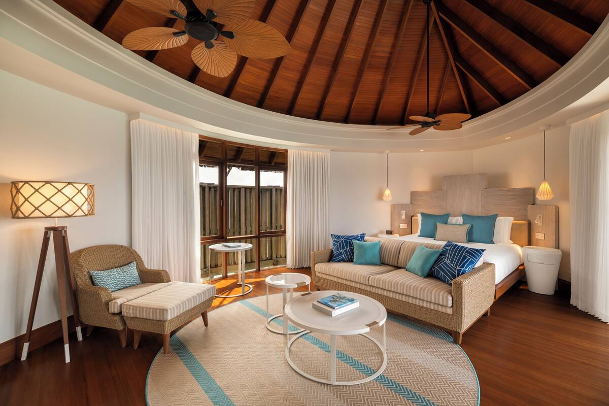 A modern hotel room with a wooden ceiling, a bed with multiple pillows, a sofa, a small round table, and a floor lamp.