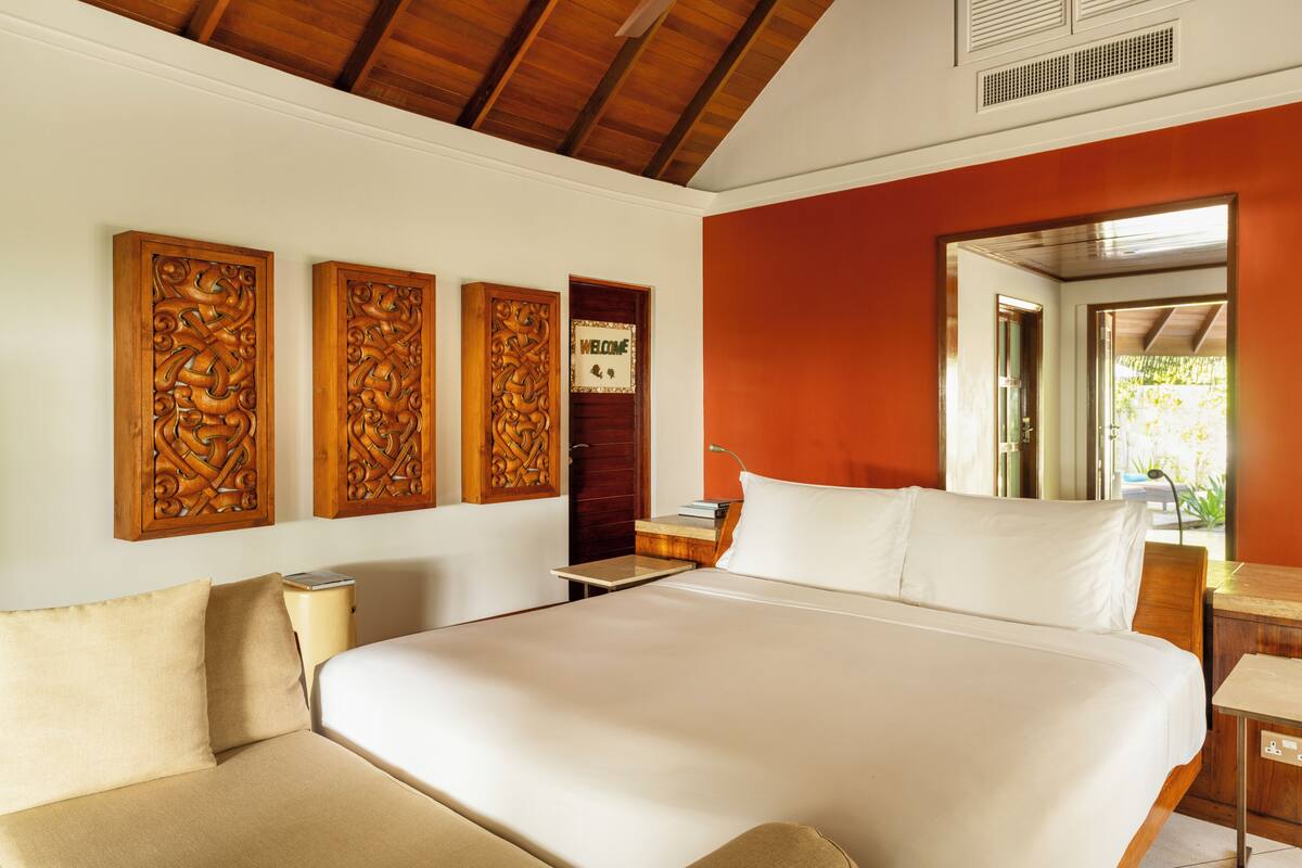 A large bed with white linens, a wooden headboard, and a nightstand with a lamp. There are three wooden carvings on the wall above the bed.