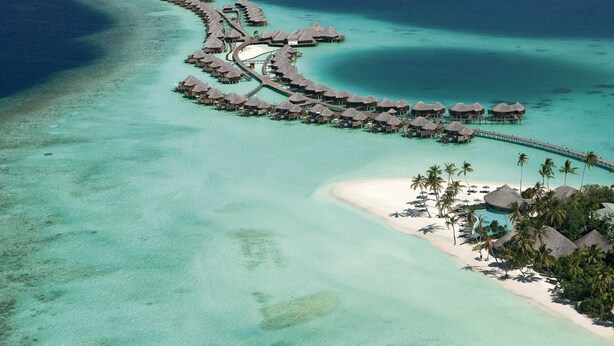 An aerial view of a resort with overwater bungalows connected by walkways, clear turquoise waters, and a sandy beach with palm trees. - Constance Halaveli (Halaveli)