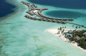 An aerial view of a resort with overwater bungalows connected by walkways, clear turquoise waters, and a sandy beach with palm trees. - Constance Halaveli (Halaveli)