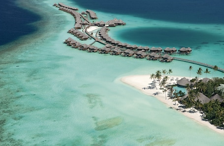 An aerial view of a resort with overwater bungalows connected by walkways, clear turquoise waters, and a sandy beach with palm trees.