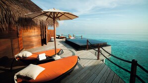 An overwater bungalow with wooden deck, lounge chairs, and a thatched roof. - Constance Halaveli (Halaveli)