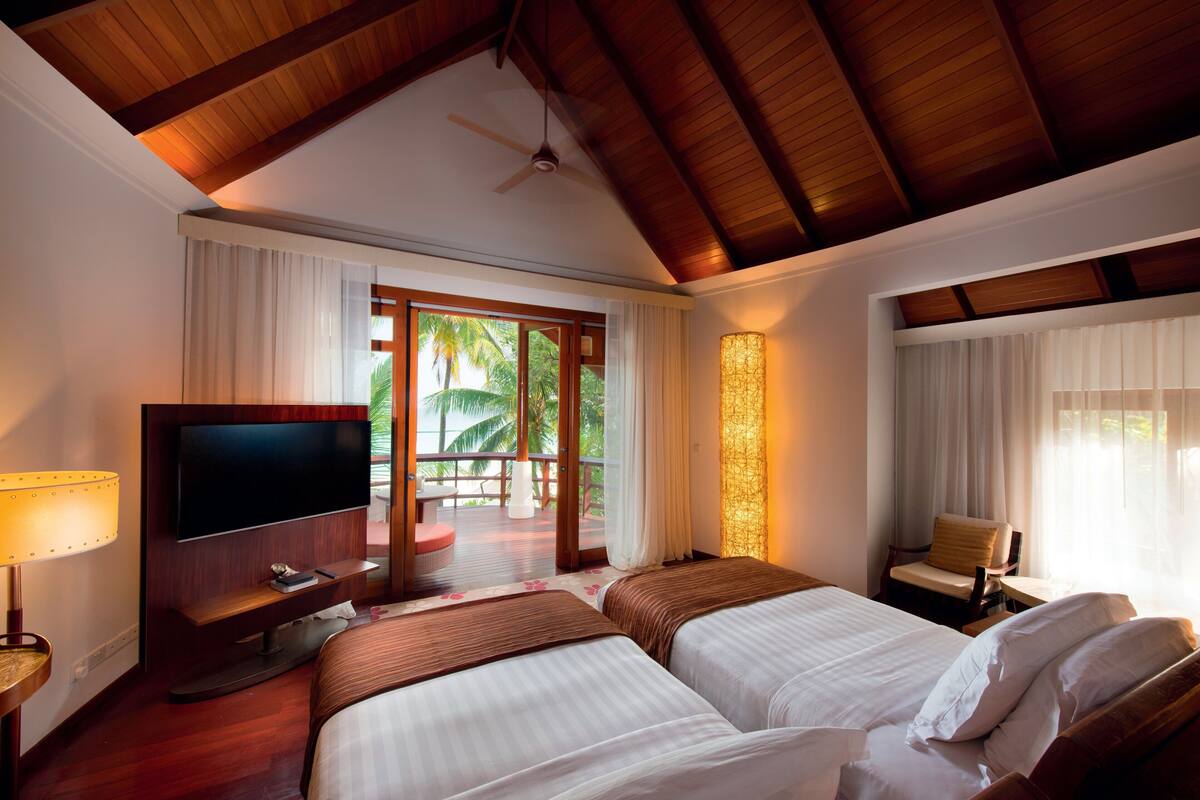 A hotel room with two beds, a TV, a chair, and a balcony with a view of palm trees.