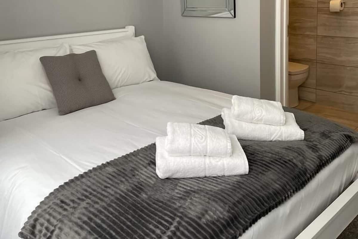 A neatly made bed with white linens, a gray throw blanket, and two folded towels. A gray pillow is on the bed, and a mirror hangs on the wall.