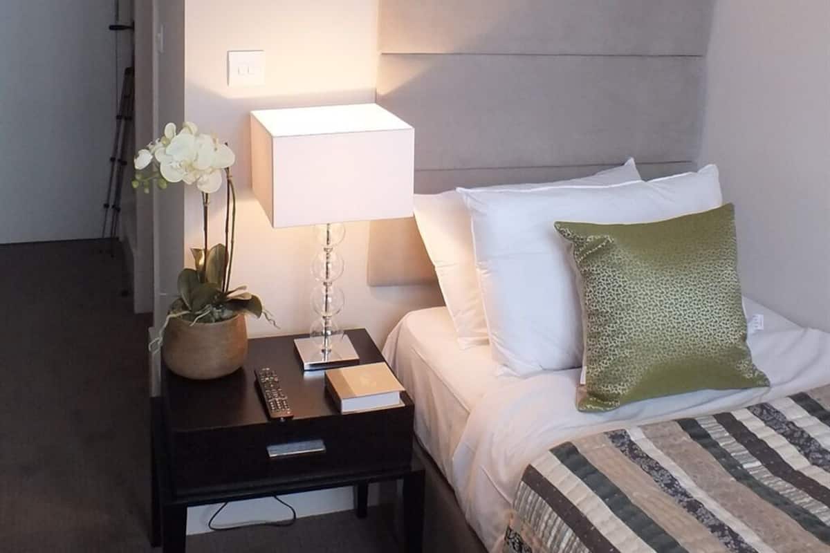 A neatly made hotel room with a bed, a bedside table with a lamp, a remote control, a book, and a potted plant.