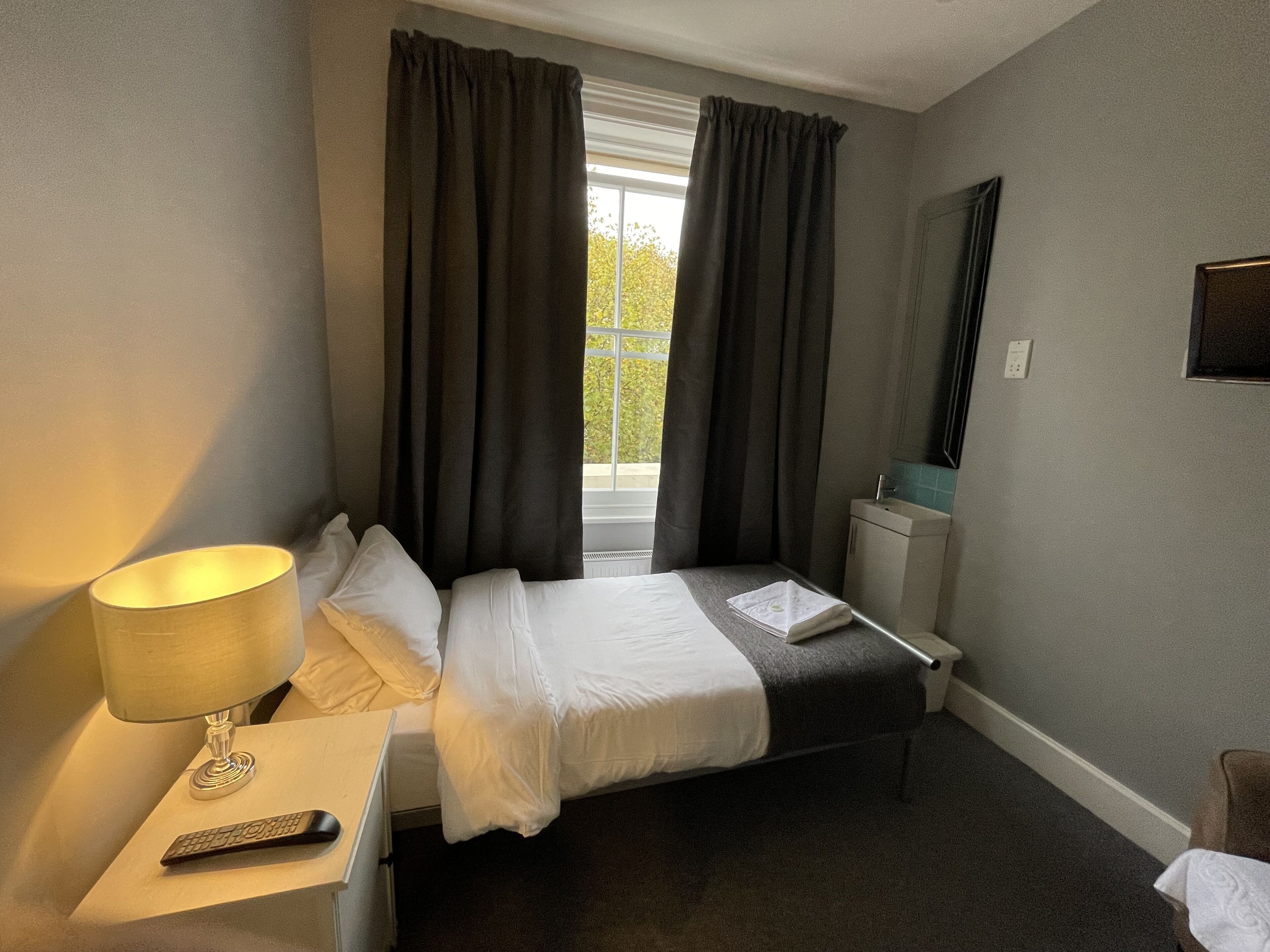 Habitación de hotel con cama, mesita de noche con lámpara, control remoto y ventana con cortinas.