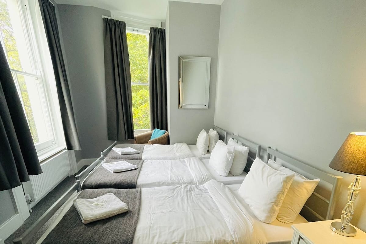 A hotel room with two beds, a bedside table with a lamp, a window with curtains, and a mirror on the wall.