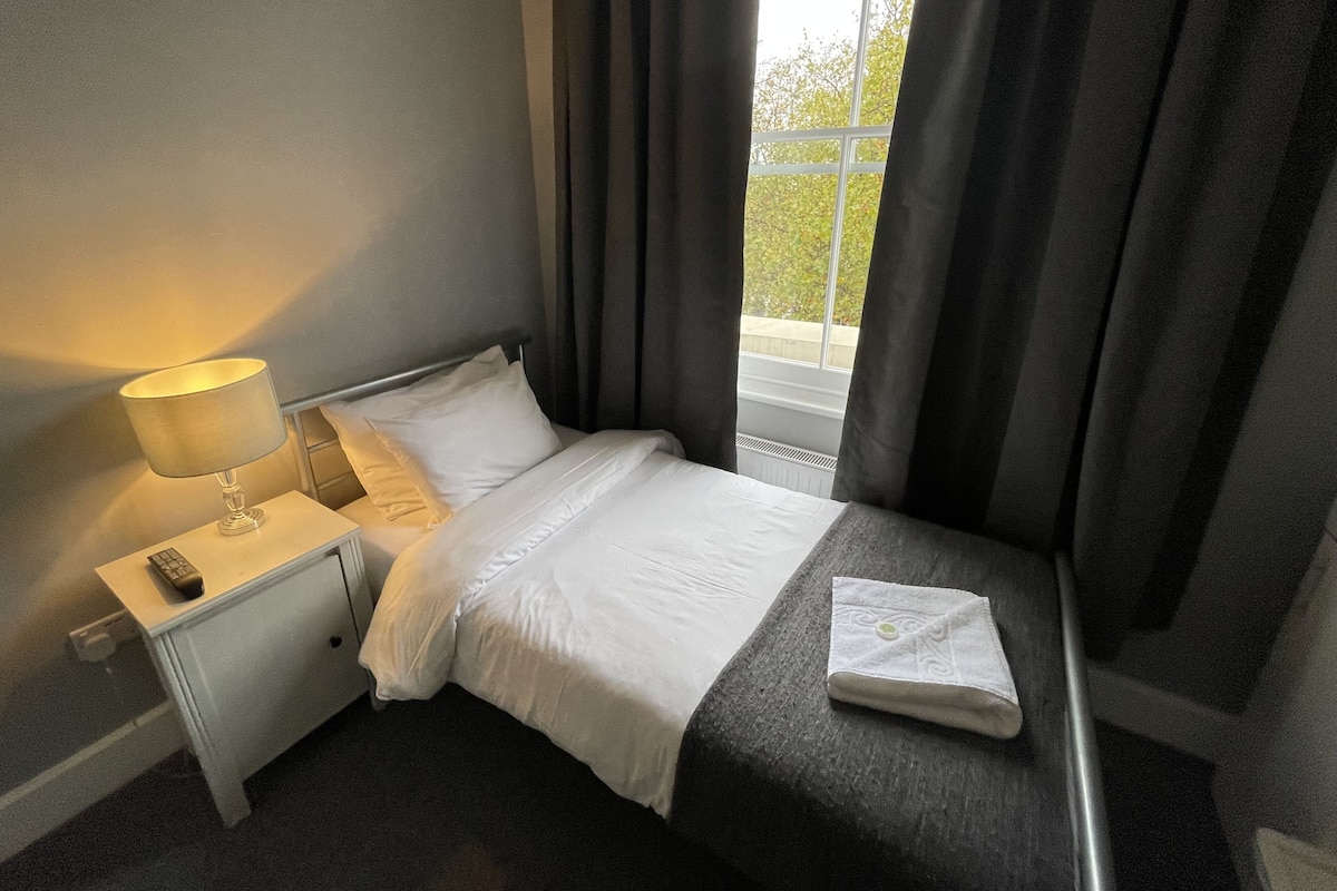 A hotel room with a bed, a bedside table with a lamp, a remote control, and a window with curtains.
